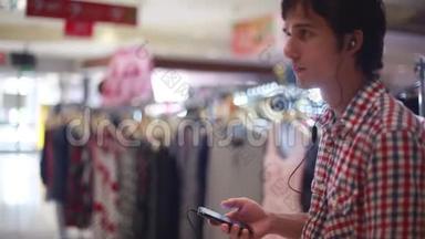 年轻的帅哥听音乐带着耳机和手机看商店。 在商场上为男人购物`